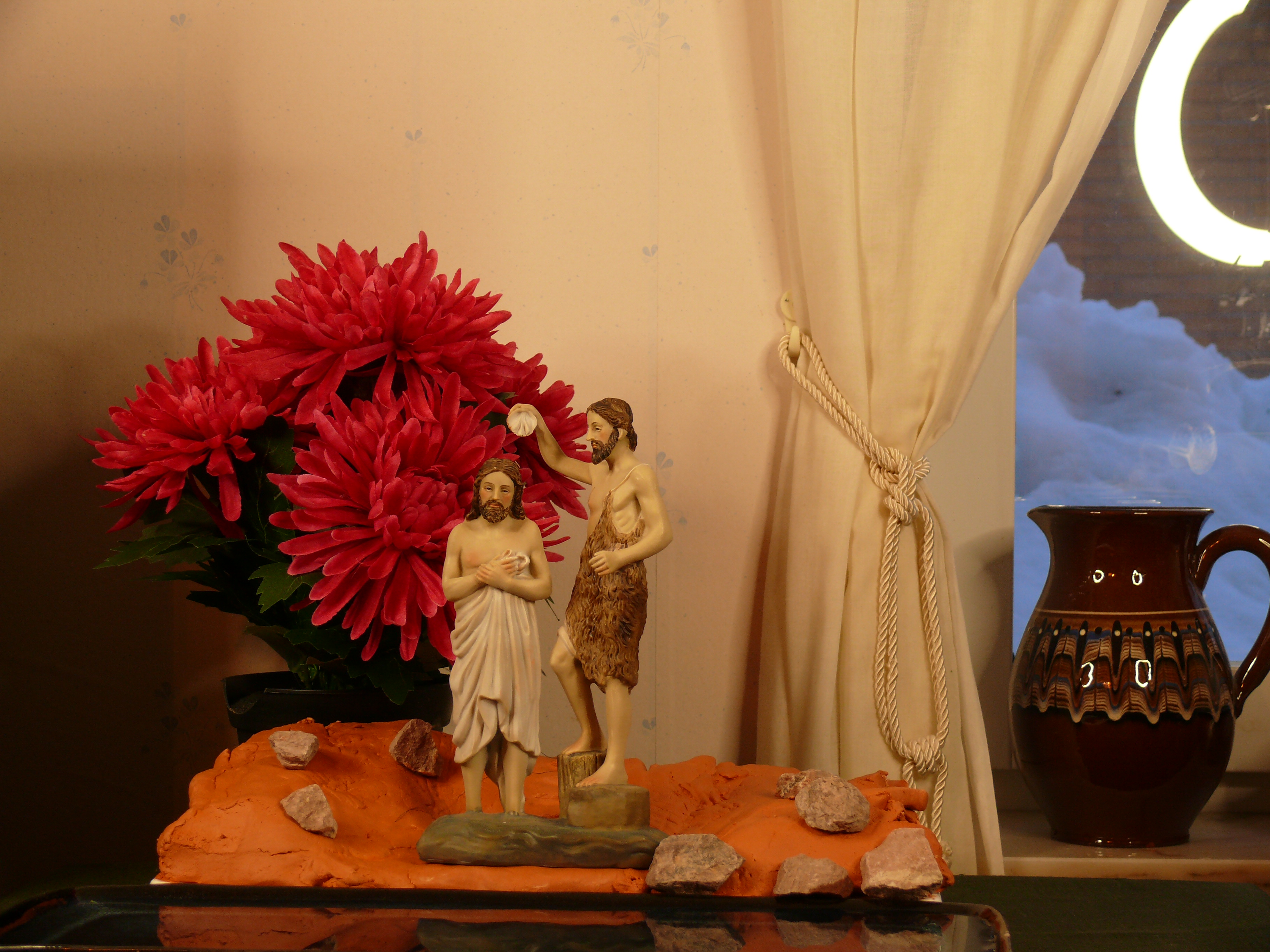 A figurine of Jesus and John the Baptist. They are standing on a clay model of  a riverbank, with a Chrysantemum in the background.